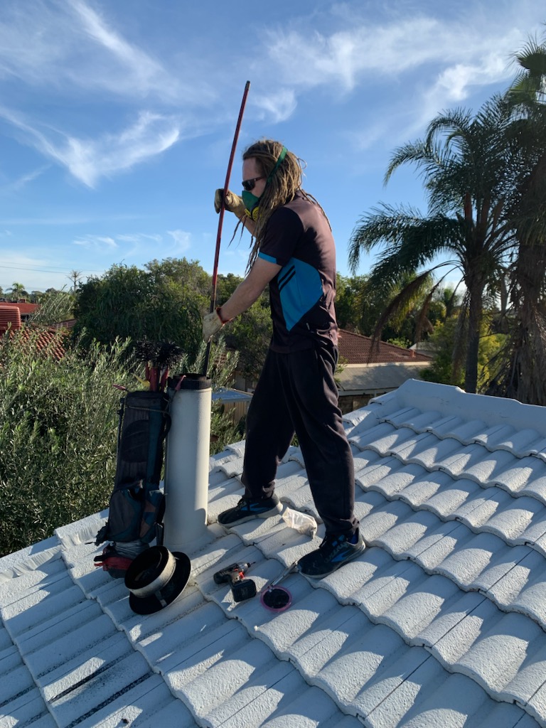 A person standing on a tiled roof, equipped with a long pole. The individual, with long hair, is wearing a black and blue shirt and a mask, appearing to be engaged in chimney cleaning work. The rooftop is surrounded by trees and a clear blue sky.