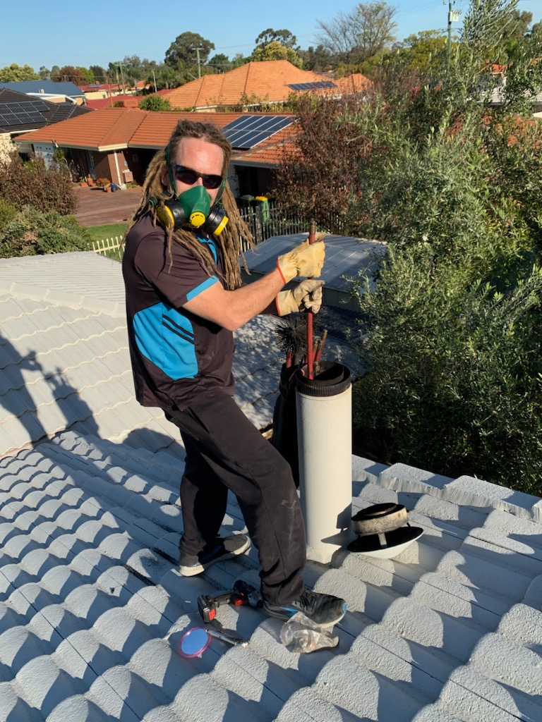 A person with long hair and dreadlocks, dressed in a black and blue work uniform, is standing on a tiled roof. They are wearing a gas mask and gloves while using a tool to clean a chimney. In the background, there are houses with solar panels and trees, indicating a suburban environment.